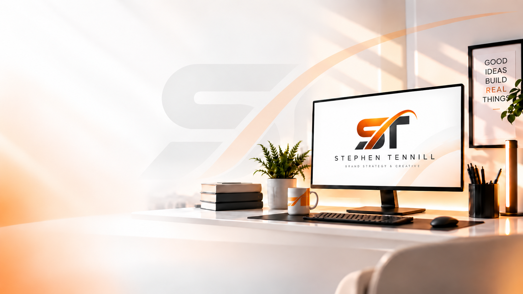Desk with a monitor showing the Stephen Tennill logo, stacked design books, a plant, and a mug.